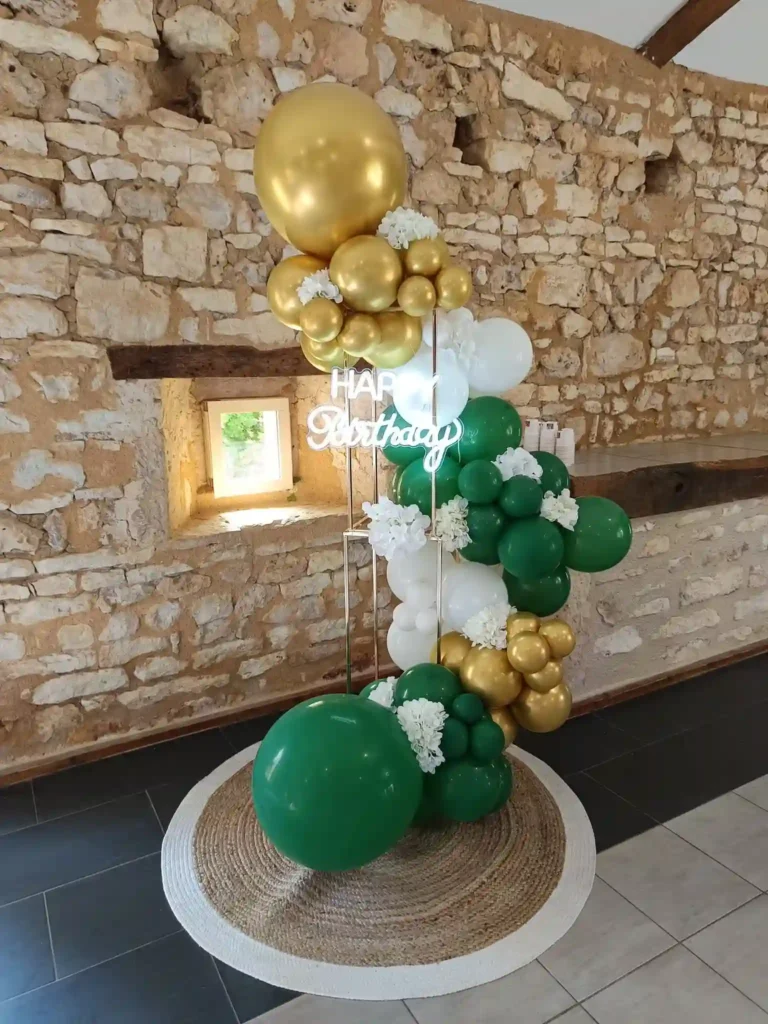 Structure ballon organique vert, blanc et or avec panneau néon « Happy Birthday » sur tapis rond en jute.